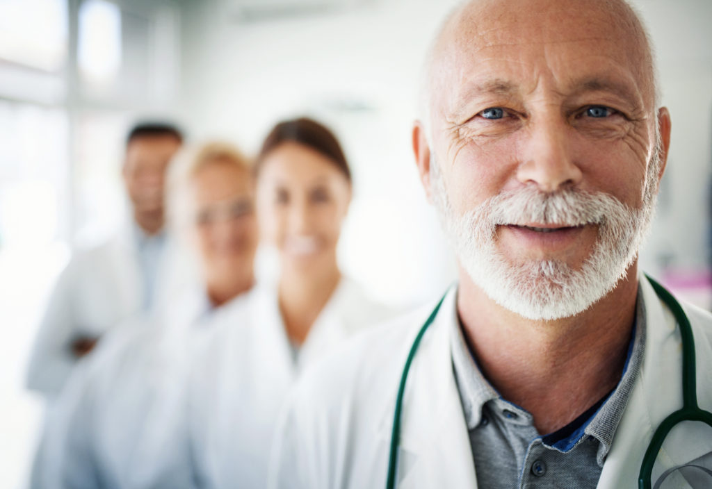 Closeup front view of group of unrecognizable doctors and nurses standing side by side and looking at the camera. Senior male doctor is in foreground, with the rest behind him and out of focus.
